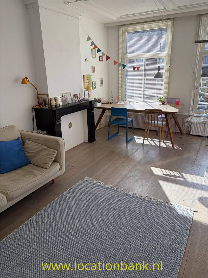 Woonkamer met een groot raam met jaloezieën, een bureau, twee stoelen, een bank en een zwarte schouw.