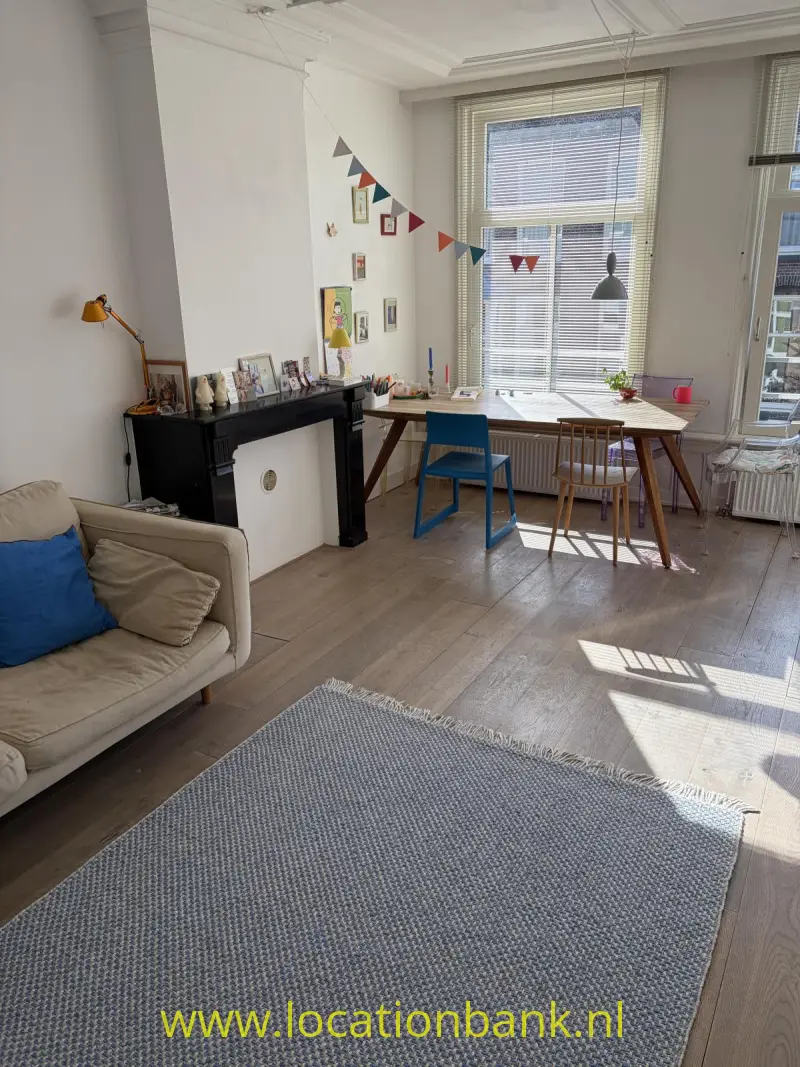 Woonkamer met een groot raam met jaloezieën, een bureau, twee stoelen, een bank en een zwarte schouw.