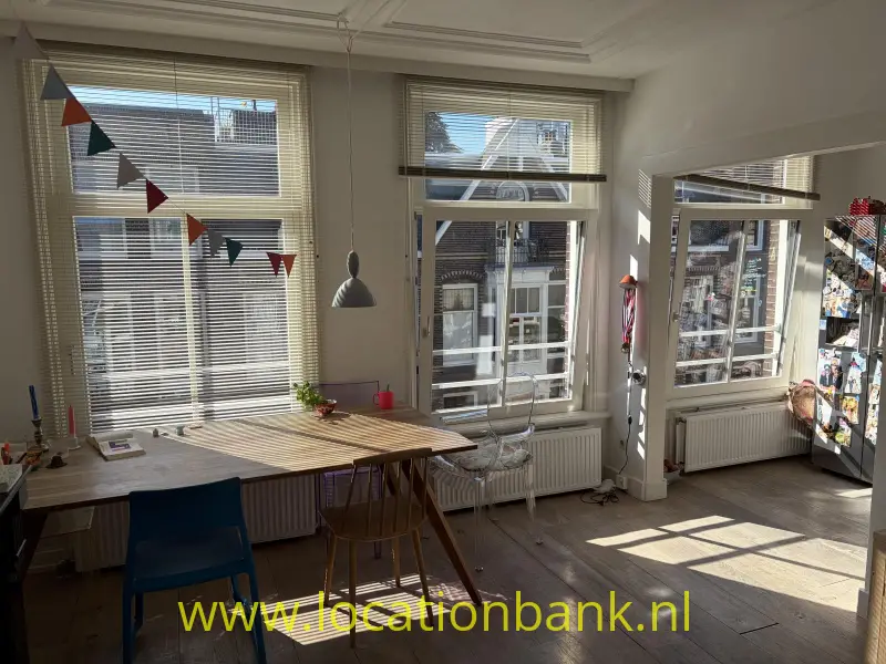 Lichte woonkamer ruimte met een houten tafel, stoelen, grote ramen en een slinger met vlaggetjes.