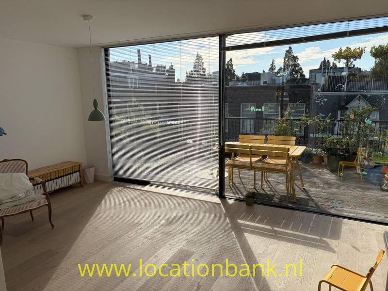 Huiskamer met grote glazen deuren met horizontale jaloezieën geven uitzicht op een terras met een gele tafel en stoelen.