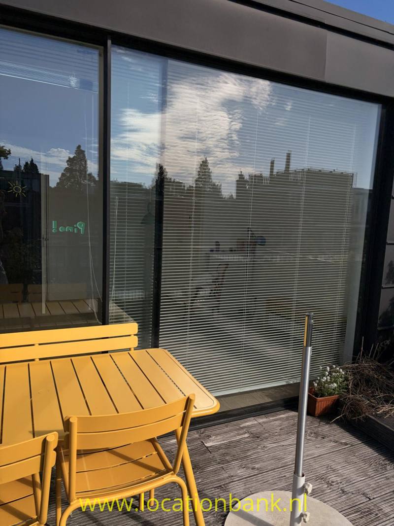 Een gele tafel met stoelen staat voor glazen deuren met horizontale jaloezieën en spiegelingen van bomen.