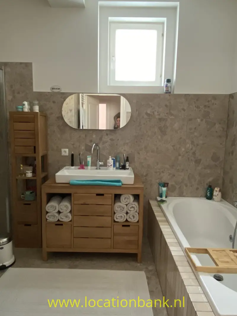 Badkamer met een houten wastafelmeubel, ronde spiegel, ligbad, beige tegelwanden en een raam.
