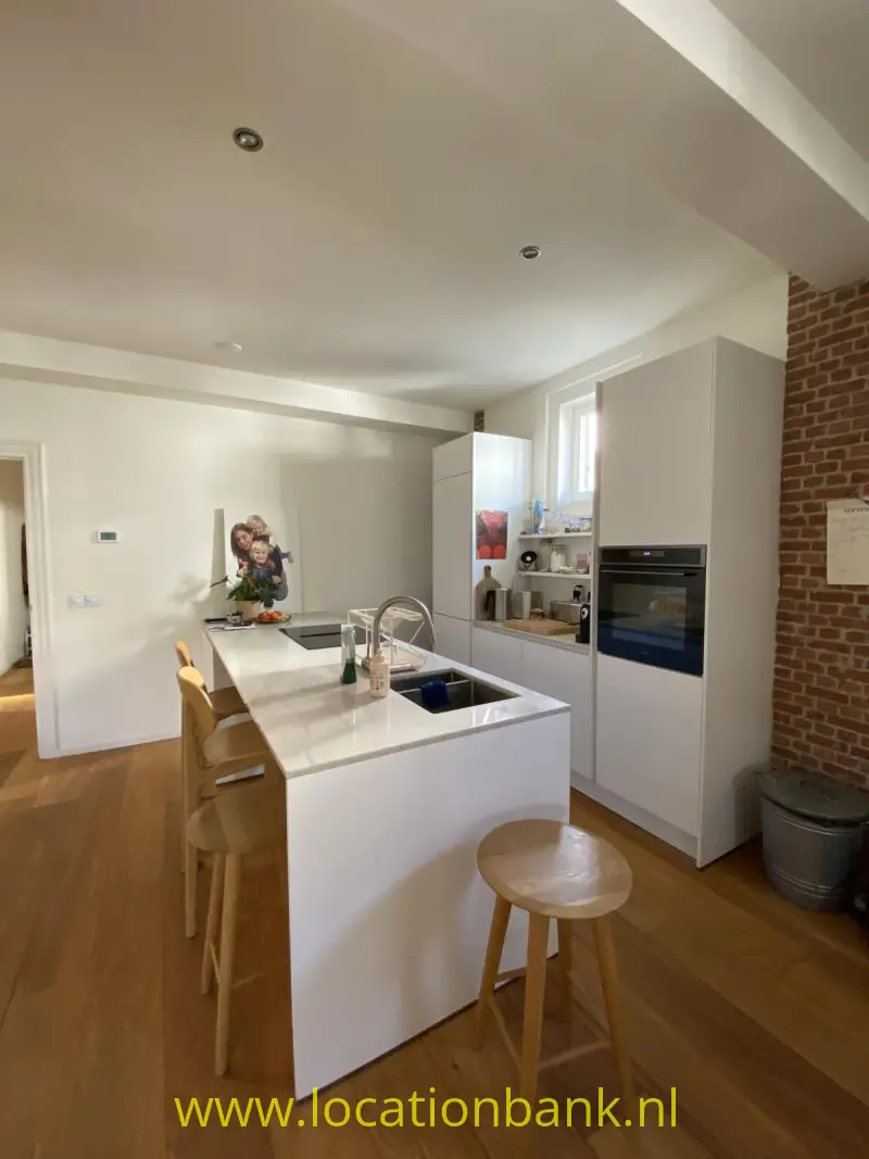 Witte keuken met een centraal kookeiland, hoge kasten, een oven en een zichtbare bakstenen muur.