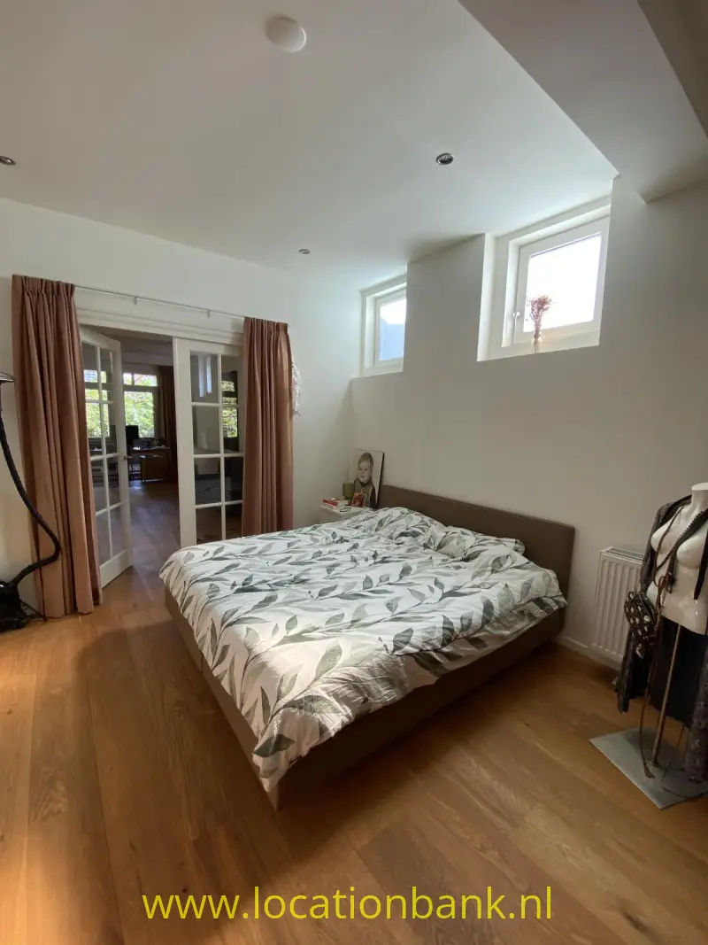 Slaapkamer met een opgemaakt tweepersoonsbed, houten vloer, gordijnen en hoge ramen.