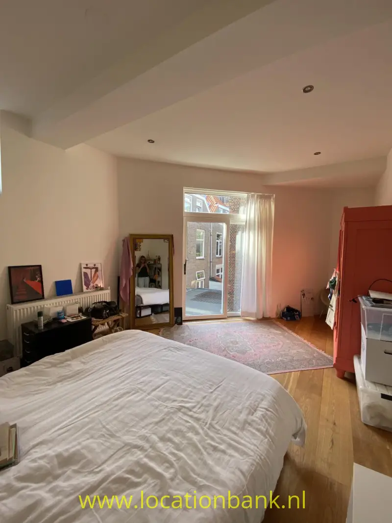 Een opgemaakt bed staat in een lichte slaapkamer met een glazen balkondeur, een vloerkleed en een kledingkast.