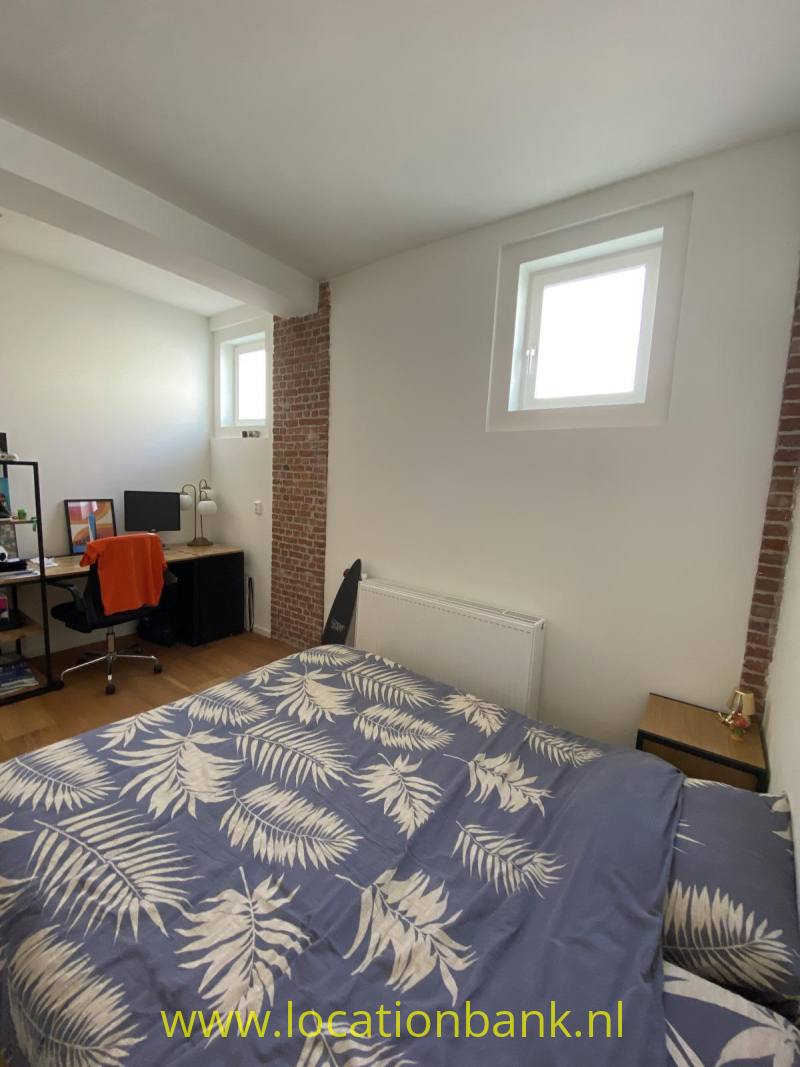 Slaapkamer met een bed met blauw beddengoed met een bladpatroon, een bureau en zichtbaar baksteenwerk.
