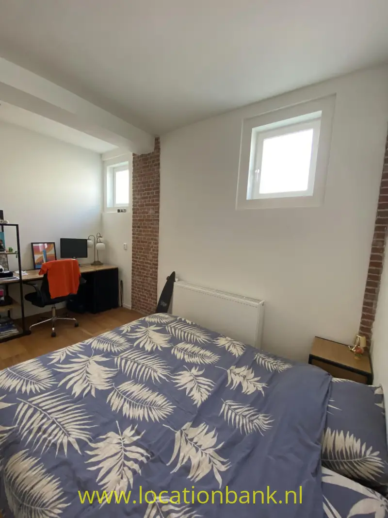 Slaapkamer met een bed met blauw beddengoed met een bladpatroon, een bureau en zichtbaar baksteenwerk.