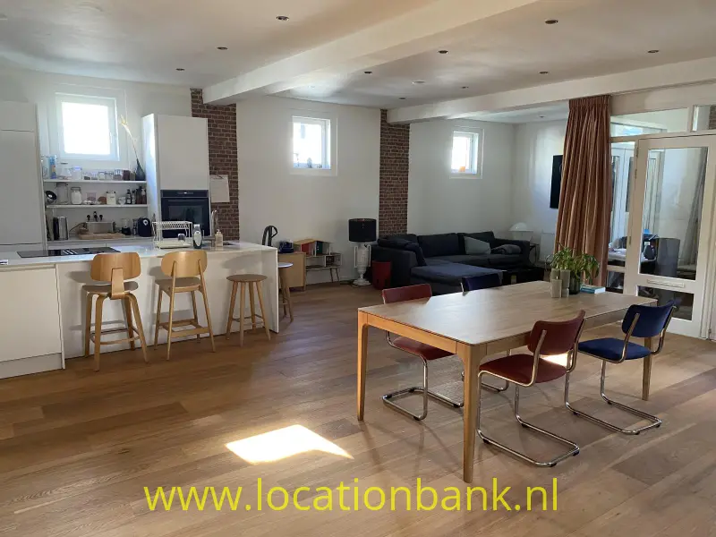 Open woonruimte met keuken, kookeiland, eettafel, zitbanken en zichtbare bakstenen zuilen.