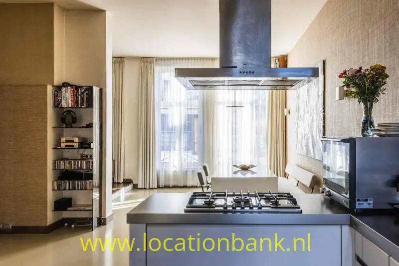 Keuken met kookeiland, gaskookplaat, afzuigkap, open boekenkast en hoge ramen.