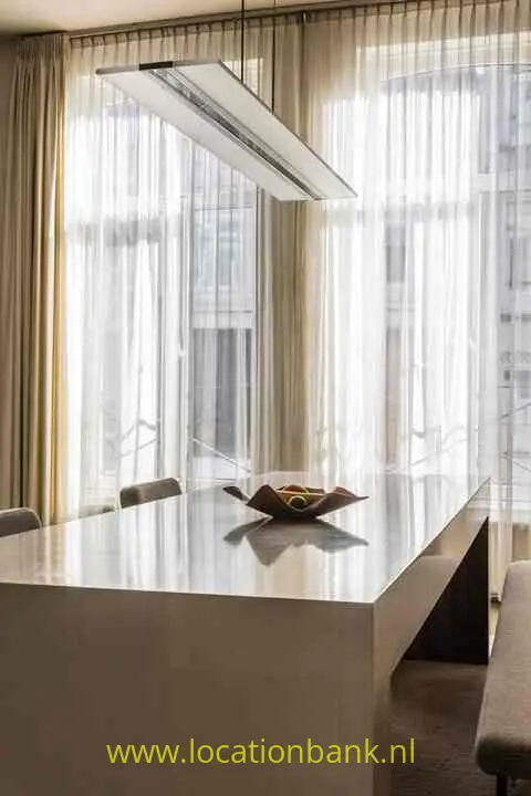 Eettafel met stoelen, een schaal en een rechthoekige hanglamp voor hoge ramen met vitrage.