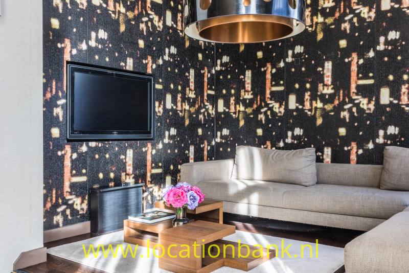 Woonkamer met een beige hoekbank, een donkere wand met lichtpatroon, een televisie en een ronde hanglamp.