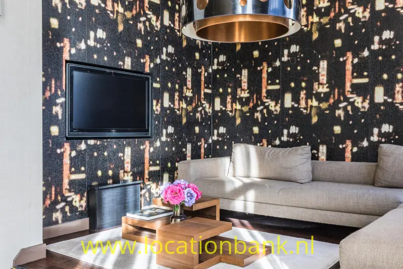 Woonkamer met een beige hoekbank, een donkere wand met lichtpatroon, een televisie en een ronde hanglamp.