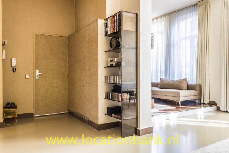 Een hal met een glazen boekenkast biedt via een open doorgang zicht op een woonkamer met hoge ramen.