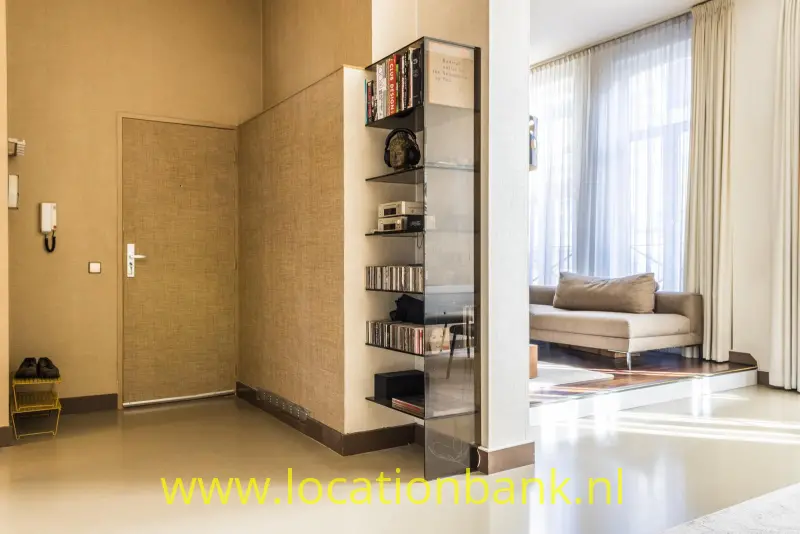 Een hal met een glazen boekenkast biedt via een open doorgang zicht op een woonkamer met hoge ramen.