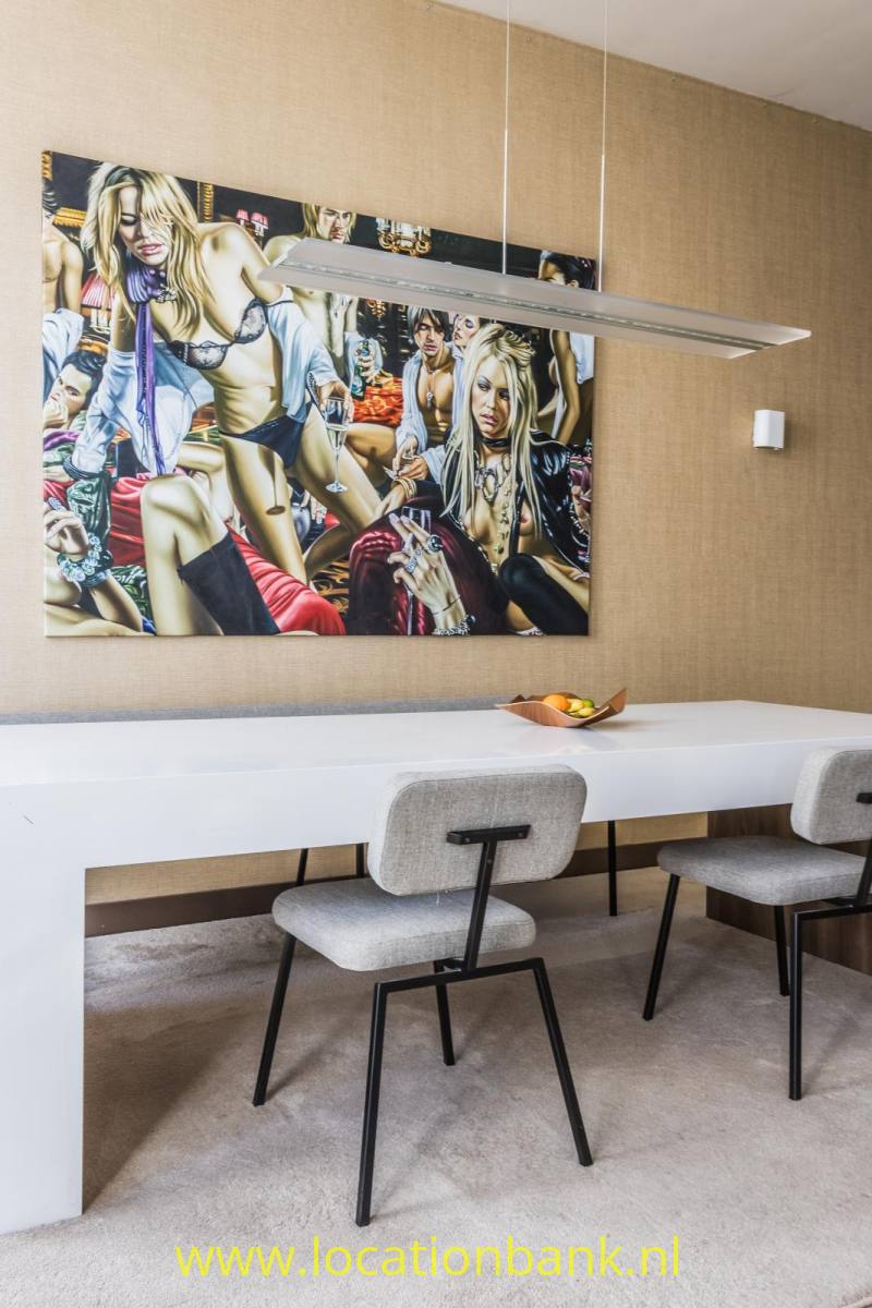 Kleurrijke wandkunst hangt boven een lange witte tafel met stoelen en een langwerpige hanglamp.