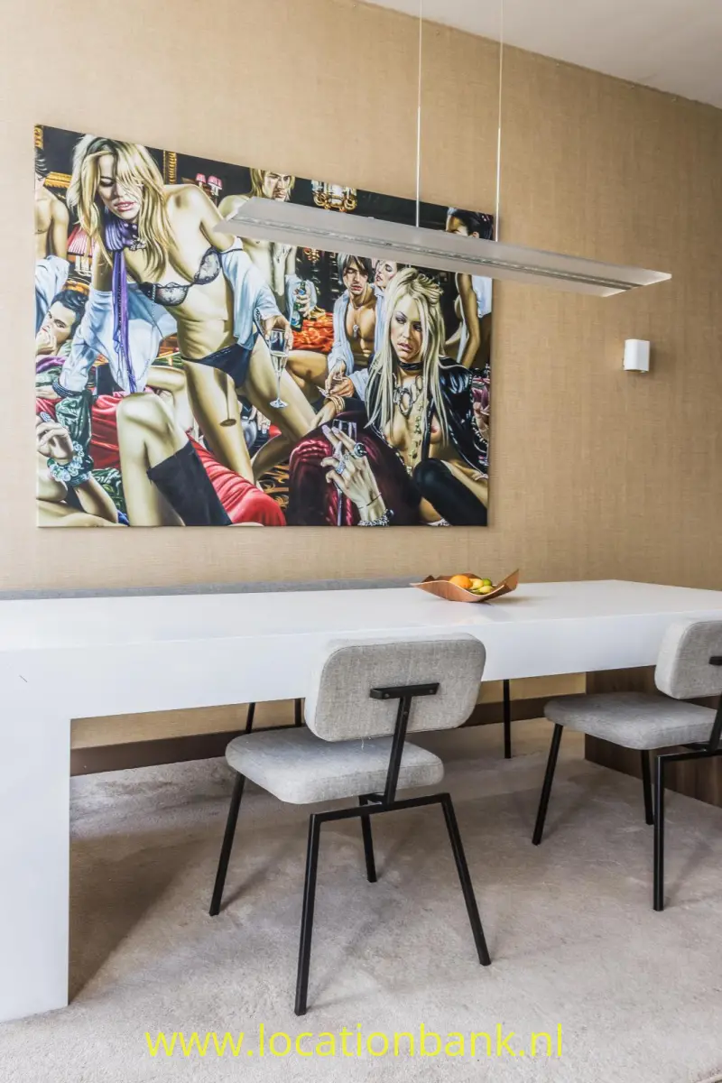 Kleurrijke wandkunst hangt boven een lange witte tafel met stoelen en een langwerpige hanglamp.