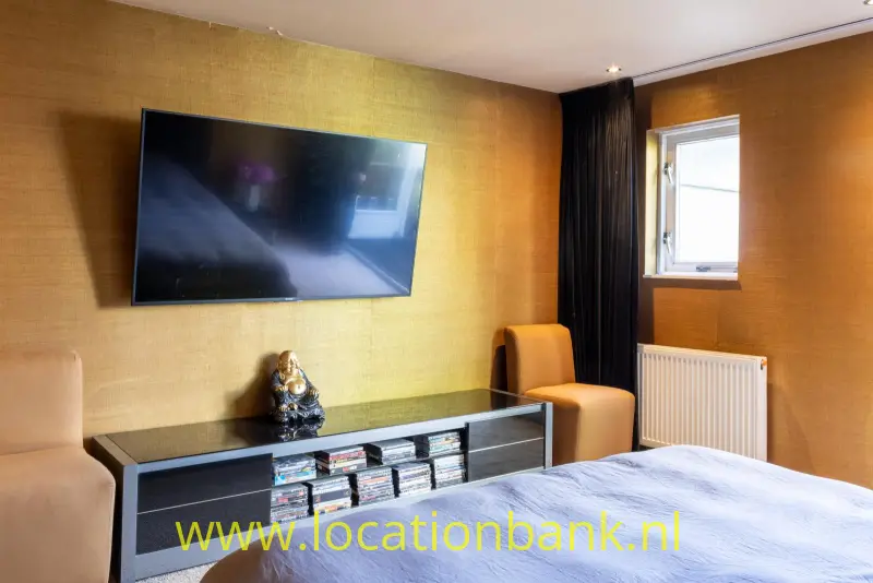 Kamer met een bed, een wandtelevisie, een zwart dressoir, een fauteuil en een raam met radiator.