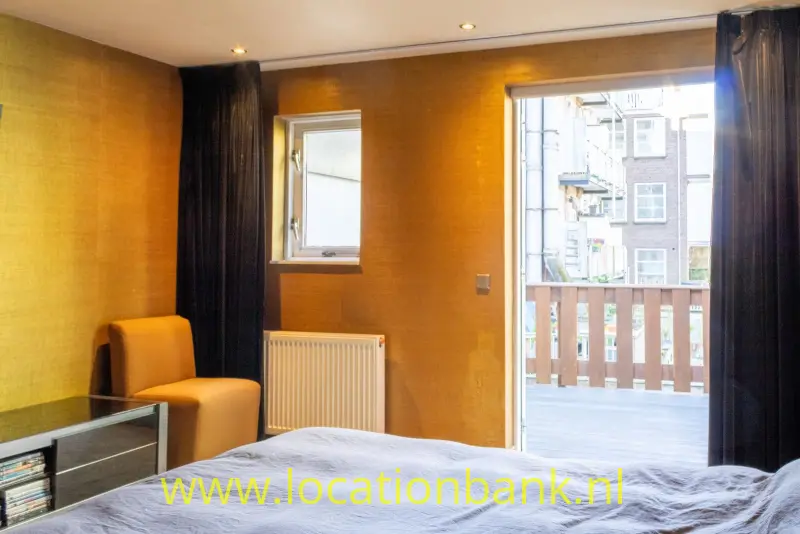 Slaapkamer met een bed, okerkleurige wanden, een fauteuil, radiator en geopende deuren naar een balkon.