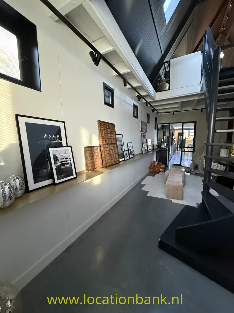 Lange binnenruimte met ingelijste kunstwerken, een open trap en dakramen boven een donkere vloer.