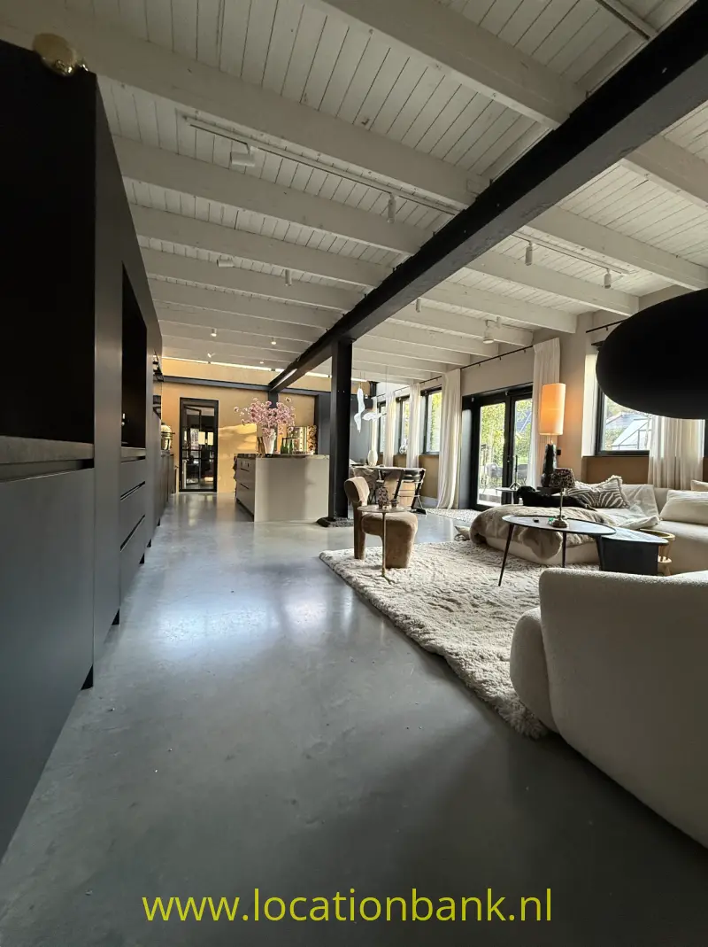 Open leefruimte met een wit balkenplafond, donkere draagbalk, zithoek, vloerkleed en grote ramen.