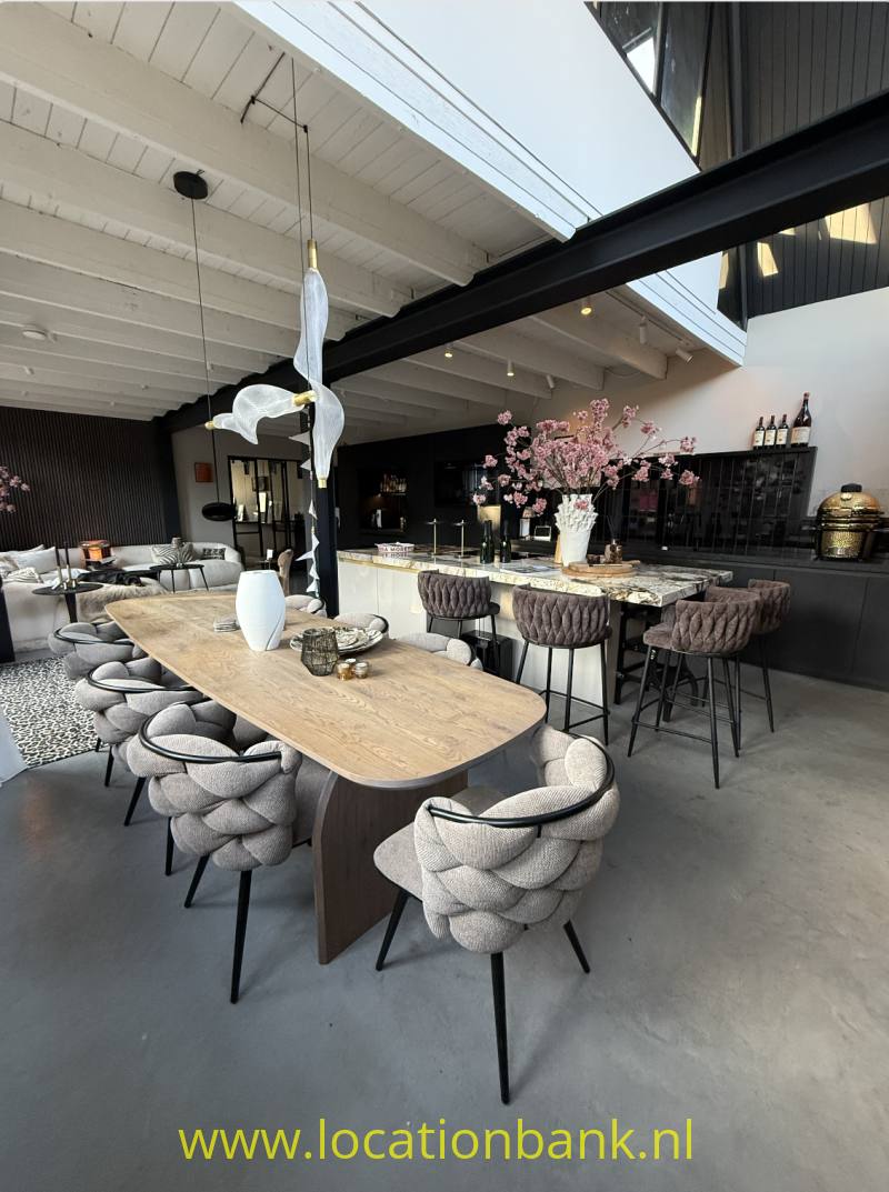 Lange houten eettafel met gestoffeerde stoelen staat in een open interieur naast een bar met bloesemtakken.