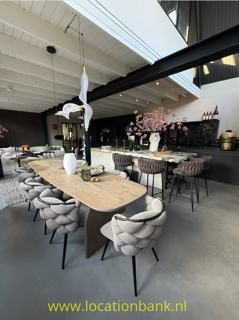 Lange houten eettafel met gestoffeerde stoelen staat in een open interieur naast een bar met bloesemtakken.