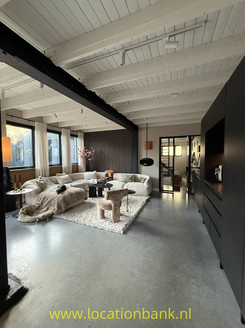 Woonkamer met een lichtgekleurde hoekbank op een licht vloerkleed, zichtbare plafondbalken en donkere kasten.