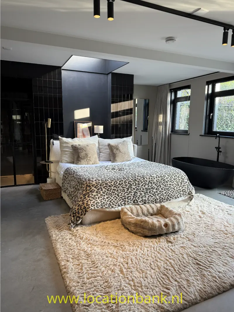 Een opgemaakt bed met een luipaardprint dekbedovertrek staat voor een zwart betegelde wand op een beige vloerkleed.