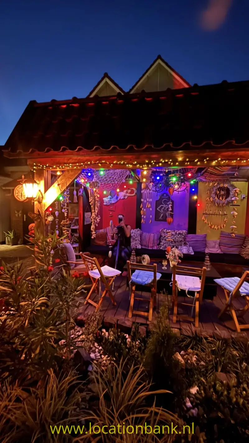 Kleurrijk verlicht terras met houten tafels en stoelen onder een donker pannendak, omringd door beplanting.