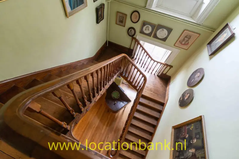 Houten trap met een gedetailleerde balustrade, ingelijste kunstwerken aan de muren en dakramen bovenaan.