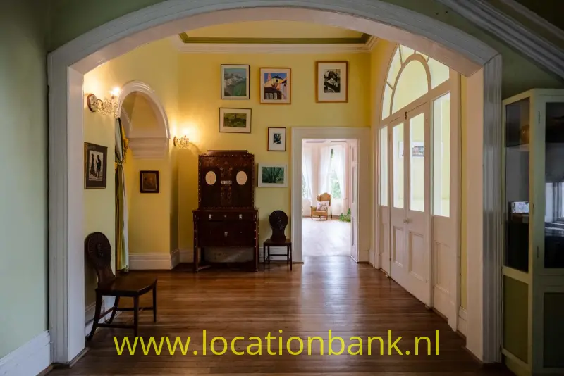 Entree Landhuis met Gele hal, houten vloer, boogdoorgang, ingelijste kunstwerken en hoge glazen deuren.
