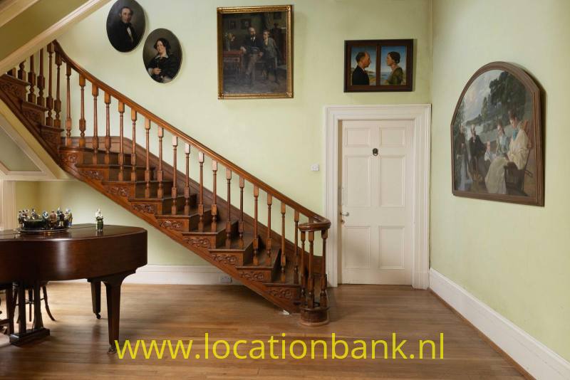Houten trap met gedetailleerde balustrade naast een vleugel, schilderijen en portretten in een interieur.