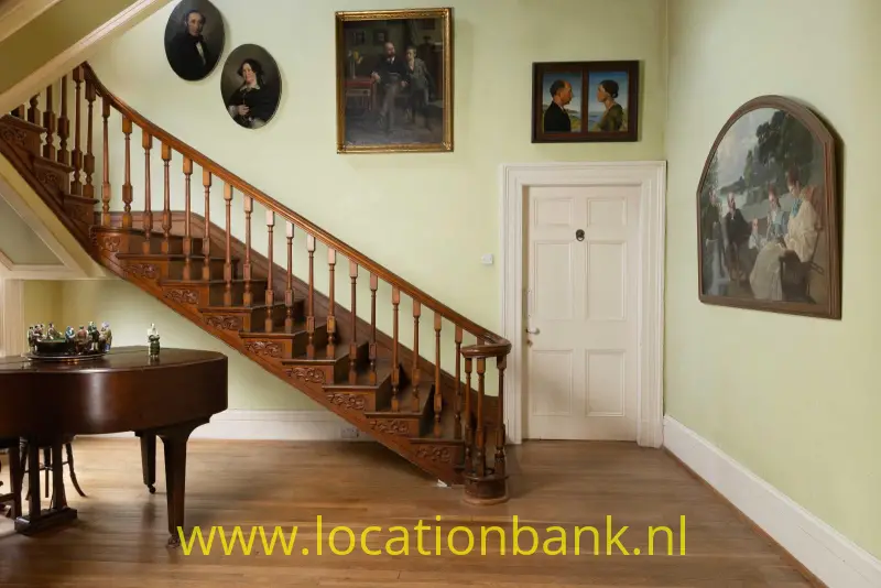 Houten trap met gedetailleerde balustrade naast een vleugel, schilderijen en portretten in een interieur.