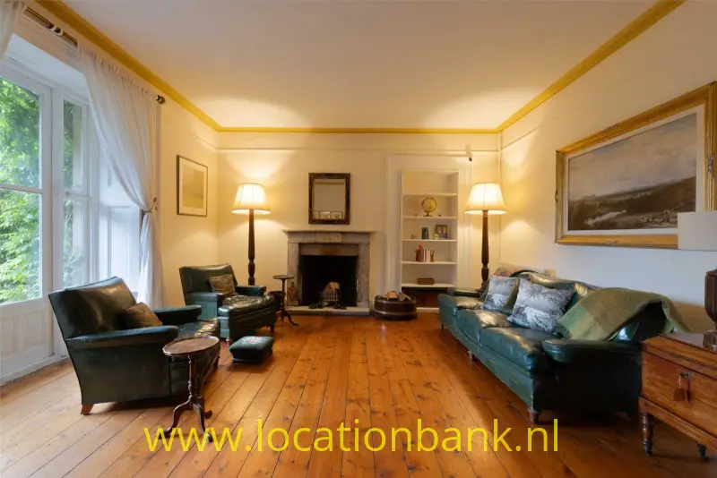 Woonkamer met een groene leren bank, fauteuils, een open haard en een houten vloer.