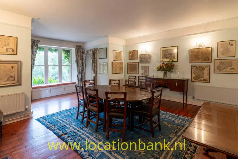 Grote houten eettafel met stoelen staat op een blauw kleed, omringd door ingelijste kaarten en ramen.