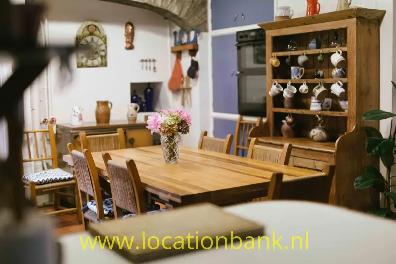 Een houten eettafel met een vaas bloemen staat voor een open kast met hangende kopjes.