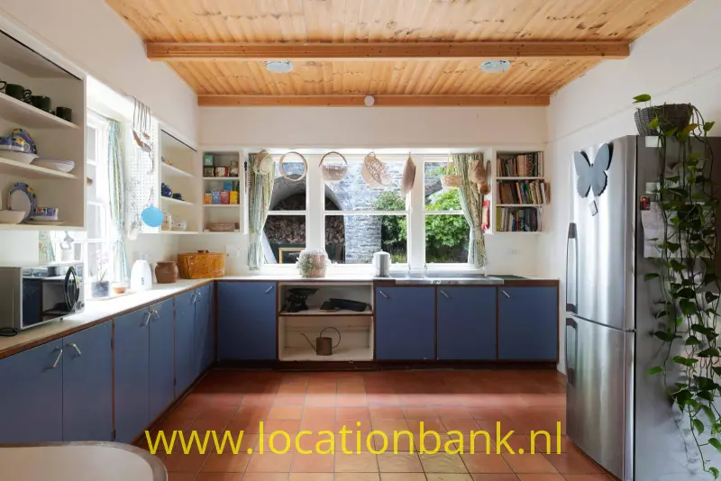 Keuken met blauwe onderkasten, een houten plafond, terracotta vloertegels en een groot raam boven het werkblad.