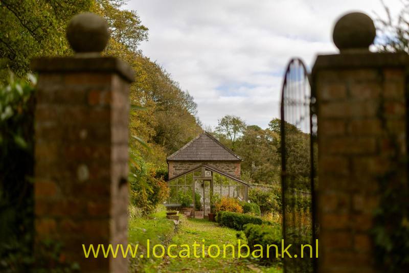 Bakstenen toegangspijlers met ronde bollen flankeren een open smeedijzeren hek en een tuinpad naar een klein houten vakwerkgebouw.