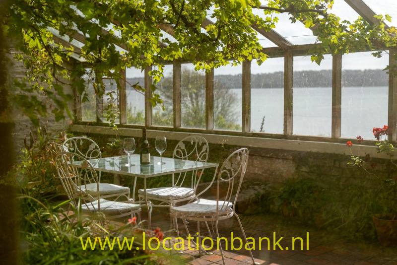 Overdekt terras in druiven kas met glazen wanden, een metalen tafel en stoelen, omgeven door groen aan het water.
