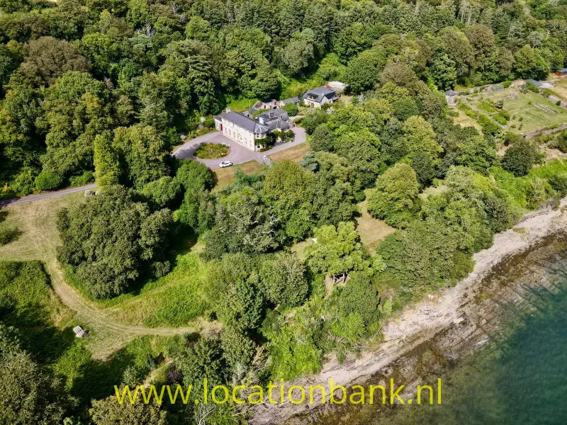 Een groot glandhuis aan zee met bijgebouwen ligt tussen dichte bomen aan een rotsachtige waterkant.