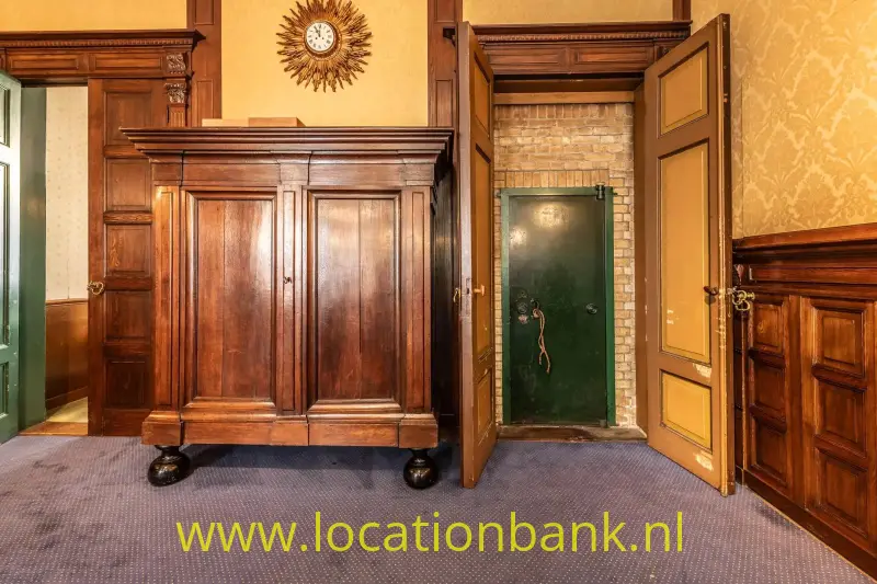 Een hoge houten kast staat onder een wandklok, naast een geopende houten deur en een groene deur in een bakstenen wand.
