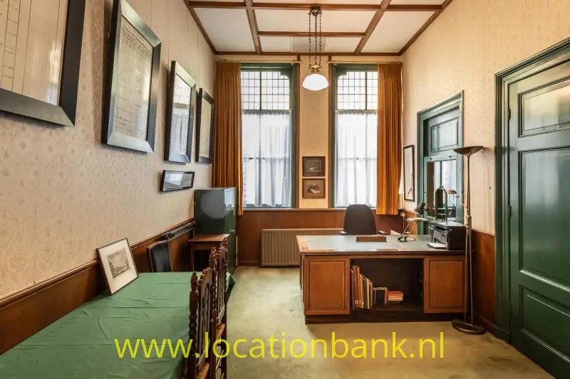 Een houten bureau staat tussen hoge ramen,  en een donkergroene deur.