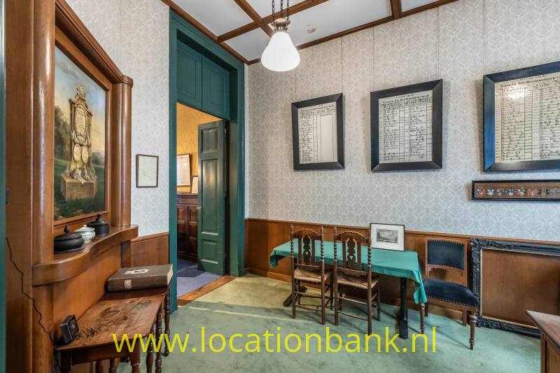 Interieur met een houten kast, groene deur, ingelijste documenten en een tafel met stoelen.