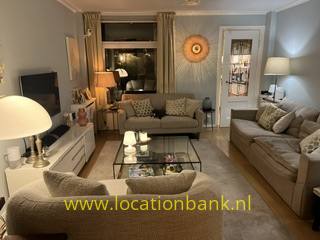 Woonkamer met twee beige banken, een glazen salontafel, tafellampen en een rond wandornament.