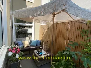 Een zithoek op een terras onder een parasol met franjes, met een houten schutting en bloeiende planten.