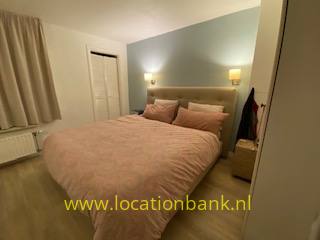 Een opgemaakt tweepersoonsbed met een roze sprei staat tegen een blauwgrijze muur, met nachtkastjes en wandlampen.