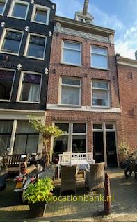 Bakstenen gevel met hoge ramen, een klein terras met stoelen en planten, en een fiets ervoor.