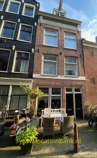 Bakstenen gevel met hoge ramen, een klein terras met stoelen en planten, en een fiets ervoor.