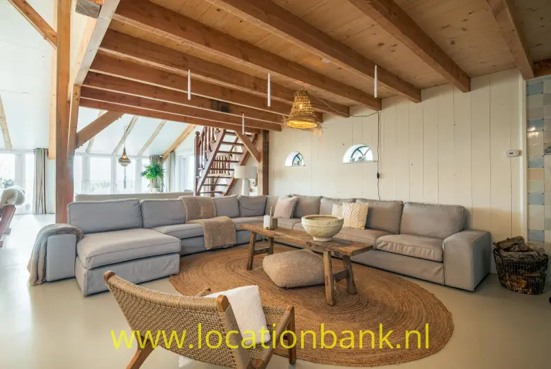 Grote grijze hoekbank onder zichtbare houten balken, met een rond gevlochten vloerkleed en een lage salontafel.