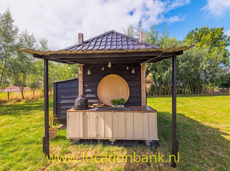Een houten kraam met een pannendak, toonbank, lichtsnoer en grote ronde wanddecoratie staat in een grasveld.